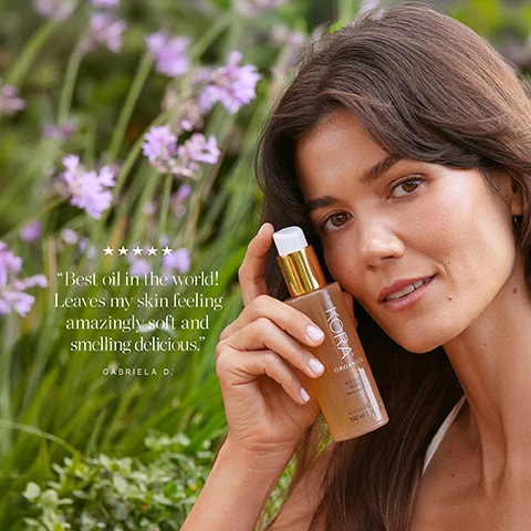 Image 1 - A woman with long dark hair holds a bottle of oil and smiles slightly, surrounded by purple flowers and greenery, with text that reads Best oil in the world! Leaves my skin feeling amazingly soft and smelling delicious. GABRIELA D.
Image 2 - A hand holding a bottle of KORA Organics Noni Glow Body Oil against a bright blue sky, with visible text that reads Best oil in the world! Leaves my skin feeling amazingly soft and smelling delicious. Gabriela D.
Image 3 - Image of two body oil products from KORA Organics, with a new packaging design on the right; the left bottle is yellow with a pump top, and the right is a tan bottle, both labeled with KORA ORGANICS and NON-GLOW BODY OIL, alongside floral illustrations and the text New Look, Same Formula.