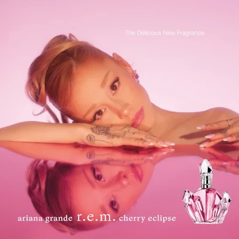 A woman with light brown hair and pink lipstick rests her head on her arms, reflecting in a pink surface, with a bottle of perfume and the text The Delicious New Fragrance and ariana grande r.e.m. cherry eclipse visible.