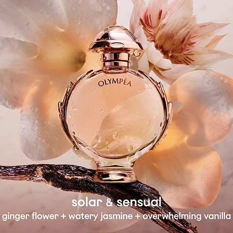 A rose gold and clear Olympéa perfume bottle with water droplets is surrounded by light flower petals and a vanilla bean pod, with text that reads OLYMPÉA solar & sensual ginger flower + watery jasmine + overwhelming vanilla.